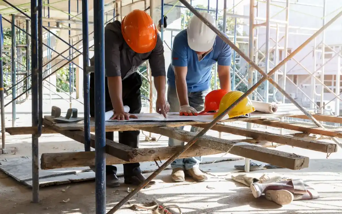 Dos técnicos de obra revisando planos sobre un tablero a pie de andamio
